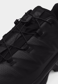 Black athletic shoe with quick-pull lacing system, textured fabric, and rubber sole designed for outdoor activities or trail running.