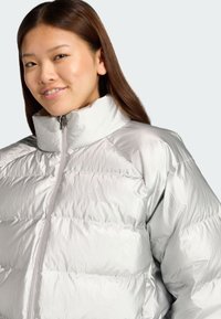 Stříbrná pufová bunda s vysokým límcem, s prošívanými částmi a zipovým zapínáním. Diskrétní logo na hrudi. Hladká, lesklá textura.