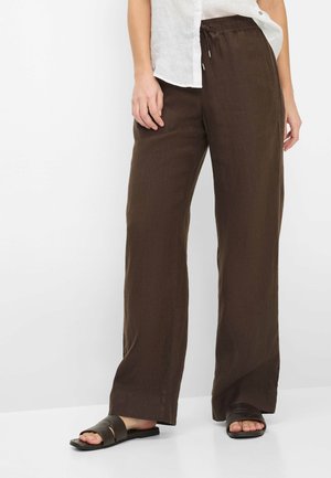 Femme portant un pantalon large marron foncé, un haut sans manches blanc et des sandales plates noires, debout devant un fond blanc.