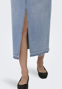 Jupe midi en denim bleu clair avec une fente à l'avant. Dotée d'un ourlet effiloché et de coutures simples. Portée avec des ballerines noires.