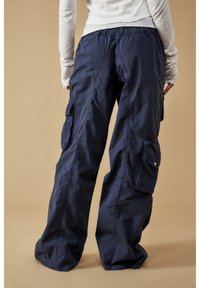 Person som bär lösa marinblå cargobyxor och en vit långärmad topp, står mot en beige bakgrund, sett bakifrån.