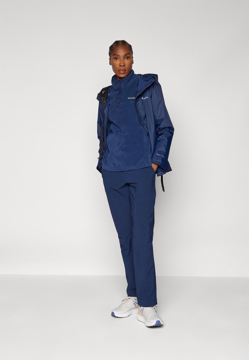 Veste imperméable bleu marine avec capuche, superposée sur un pull en polaire bleu marine et un pantalon bleu marine, portée avec des chaussures de sport blanches et grises.