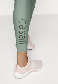 Ljusgröna leggings med "Casall"-logotyp i mörk text, kombinerat med ljusrosa träningsskor med nät- och texturdetaljer.