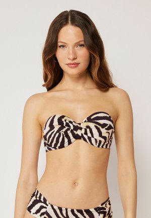 Kvinde med langt brunt hår iført en zebra-print stropløs bikini top og matchende underdel, stående mod en ensfarvet lys baggrund.