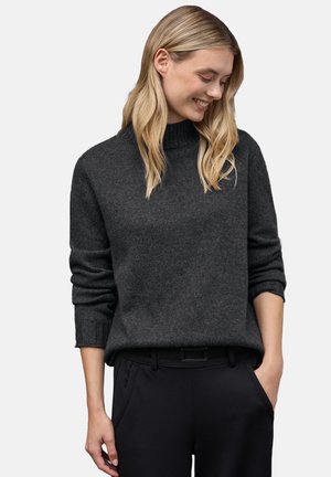 Maglione grigio antracite con collare alto, maniche arrotolate e vestibilità rilassata, abbinato a pantaloni neri. Texture morbida e design minimale.