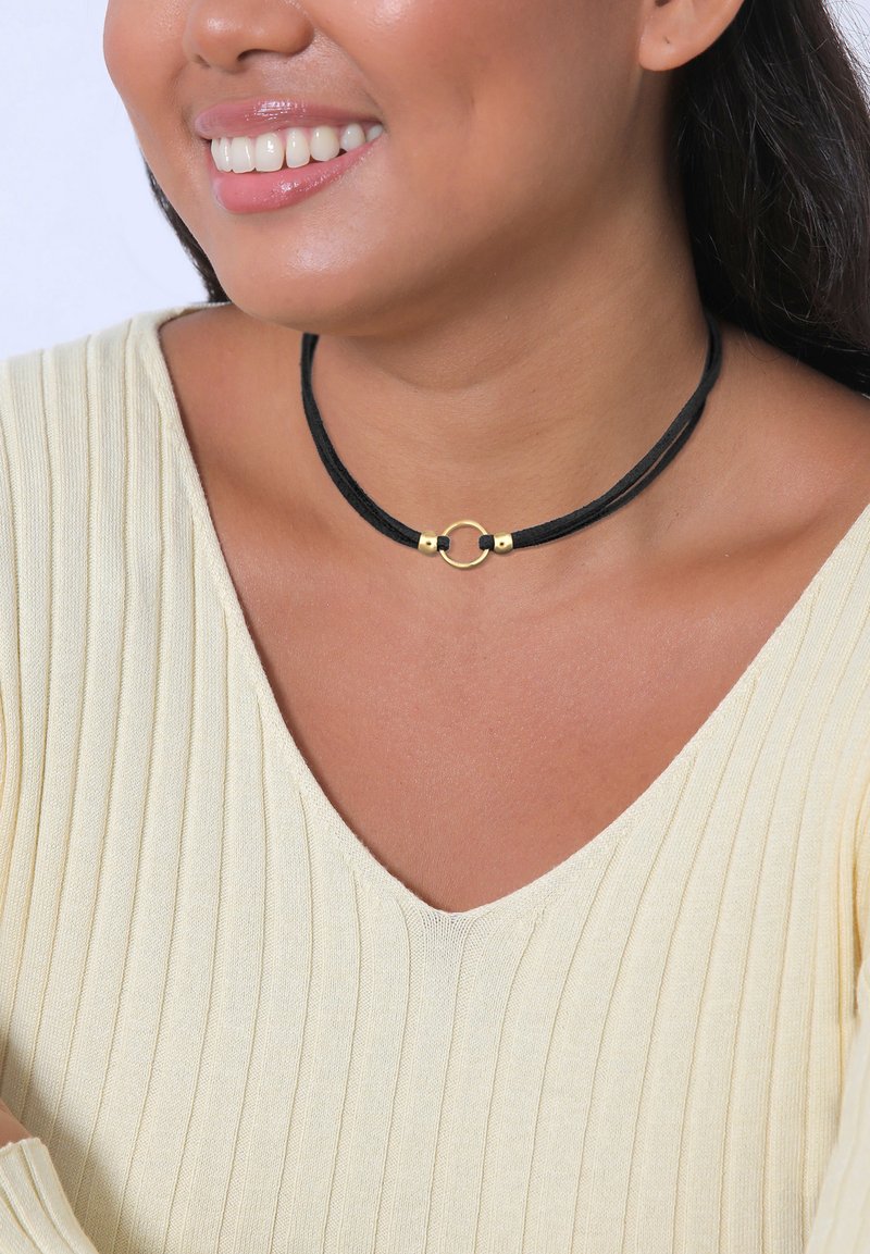 Collana choker nera composta da due fili con un anello rotondo dorato e dettagli metallici alle estremità. Indossata con un top a coste beige chiaro.