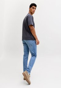 Jonge man die een donkergrijs T-shirt, lichtblauwe jeans en beige sneakers draagt, staand tegen een effen witte achtergrond.