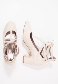 Le décolleté in suede rosa chiaro presentano lati dalle forme ritagliate, una punta arrotondata e lacci incrociati con un piccolo dettaglio dorato. Il tacco ha una forma a blocco chunky.