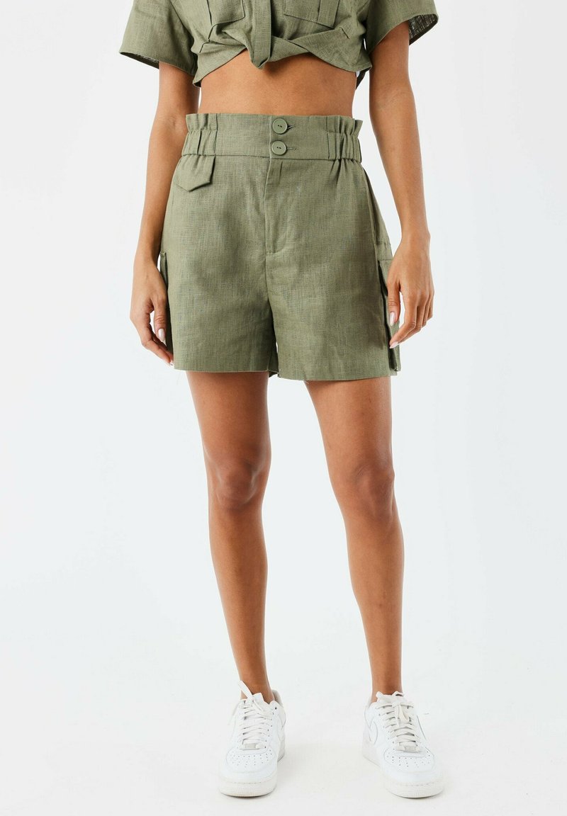 Pantalones cortos verde oliva de talle alto, con dos botones, bolsillos laterales y una tela texturada. Combinados con zapatillas blancas.