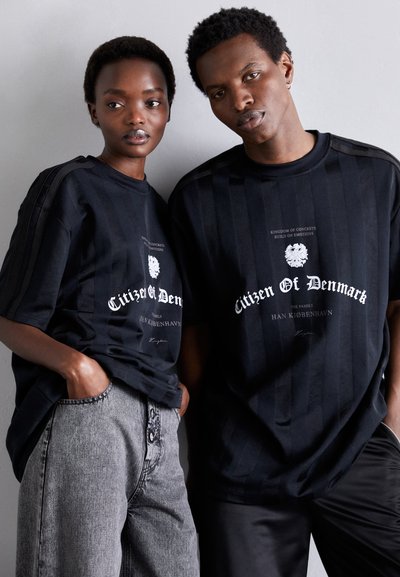 Sorte oversized t-shirts med lodret stribemønster, med hvid trykdetaljering. Båret med grå bukser, der har et lige snit.