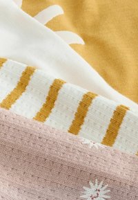 Tissus tricotés pliés en jaune moutarde, blanc avec des rayures moutarde, et rose pâle avec des motifs de soleil blancs et des détails de tissage texturé.