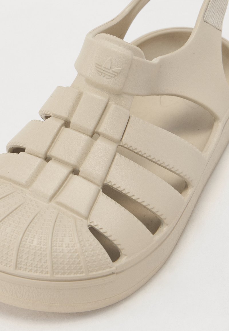 Beige Plastik-Sandale mit geschlossenem Zeh, Kreuzriemen-Design, strukturiertem Sohlenprofil und Logo auf dem oberen Riemen. Offene Seiten für Belüftung.
