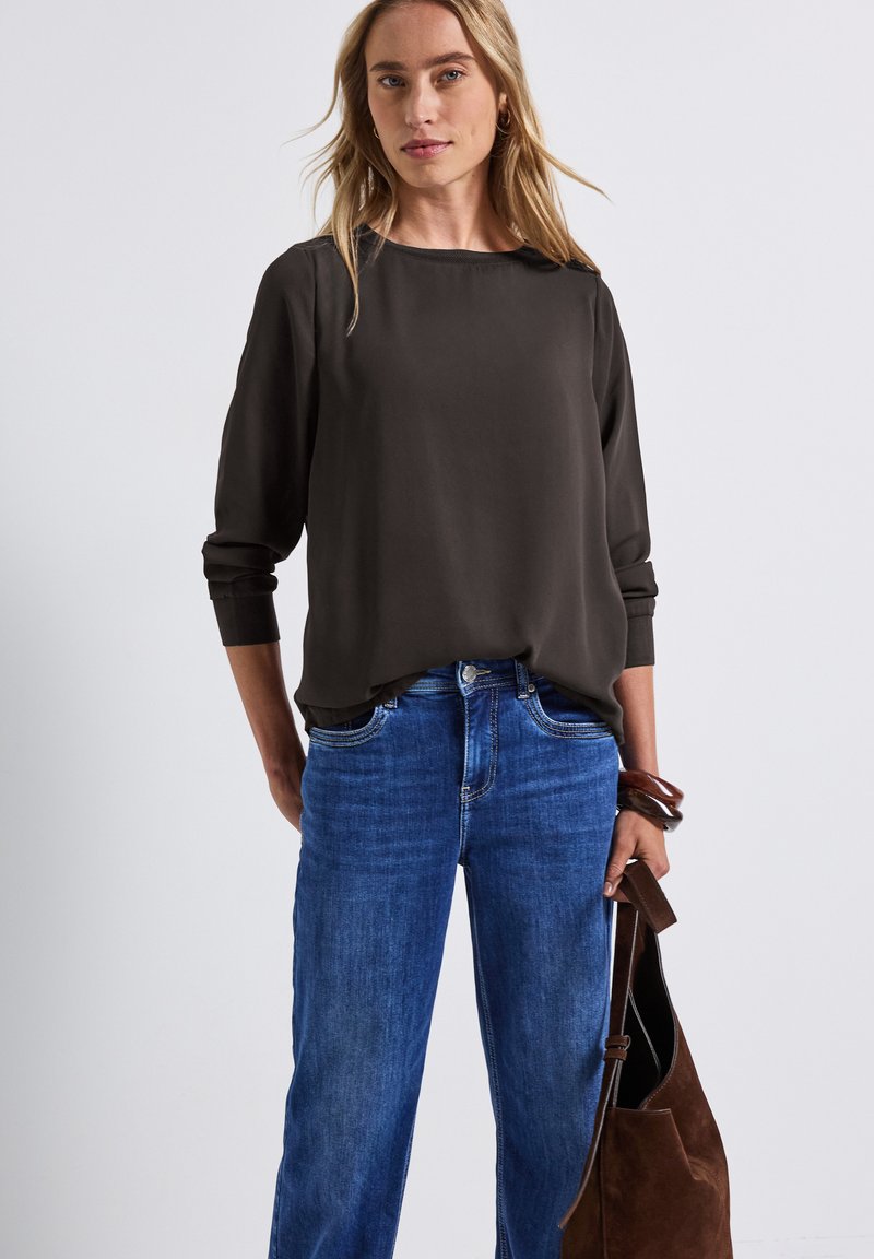 Graue Bluse mit langen Ärmeln und weitem Ausschnitt, kombiniert mit blauen Jeans und einer braunen Wildleder-Schultertasche. Weicher Stoff und entspannte Passform.
