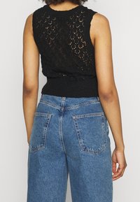 Schwarzes, gestricktes ärmelloses Oberteil mit offenem Spitzenmuster und geripptem Taillenbund, kombiniert mit hoch taillierten blauen Jeans aus Denim mit Gesäßtaschen.