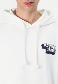 Hoodie blanc avec une capuche à cordon, comportant un logo bleu "TOMMY JEANS" sur la poitrine. Tissu doux avec une coupe décontractée.