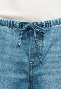 Μπλε τζιν παντελόνι με ελαστική μέση και κορδόνι δέσιμο, σε συνδυασμό με λευκό ριμπ τοπ που φαίνεται στη μέση.