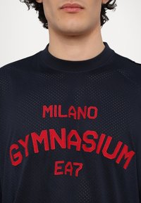 Jūras spēku acu t-krekls ar sarkano izšūto "MILANO GYMNASIUM EA7" logo. Iespējas ir apaļš kakls un īsas piedurknes ar ventilācijas detaļām.