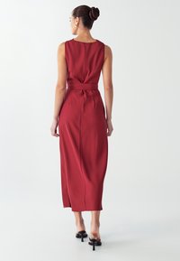 Dlouhá, bezrukávová šaty v tmavě burgundské barvě. Obsahuje opasek na zavázání, hladkou strukturu a jemný zadní zip. Délka ke středu lýtek.