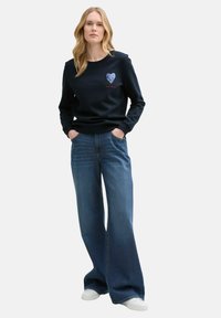Sudadera marina con diseño de corazón y logotipo, combinada con vaqueros de pierna ancha azules y zapatillas blancas. Mezcla de algodón, corte relajado.