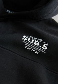 Fekete kapucnis pulóver, fehér szöveggel: "PREMIUM GOODS SUB.5 Williamsburg, Brooklyn, New York City 40.7081° N, 73.9571° W SUB5CRIBE"