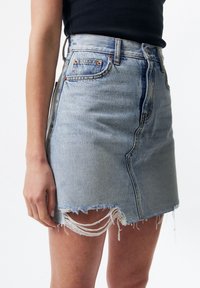 Vaaleansininen farkku-minihame, jossa on revitty helma, viisi taskua ja kuparinvärinen varustus. Pystysuora kulutustyynyksitys kankaassa.