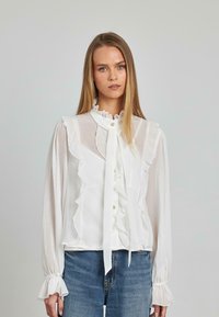 Weiße, rüschige Bluse mit langen Ärmeln, die über einen hohen Ausschnitt und eine Bindeschleife verfügt. Hergestellt aus einem durchsichtigen Material mit Knopfdetails.