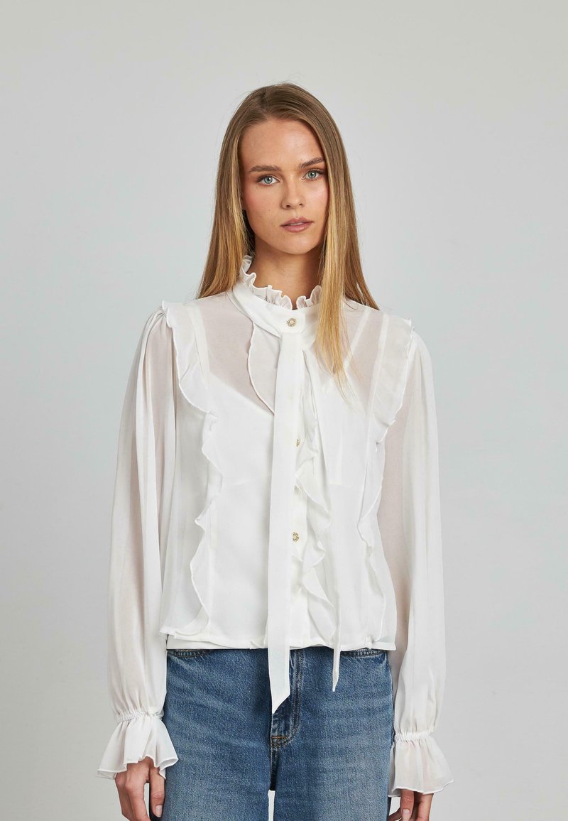 Weiße, rüschige Bluse mit langen Ärmeln, die über einen hohen Ausschnitt und eine Bindeschleife verfügt. Hergestellt aus einem durchsichtigen Material mit Knopfdetails.