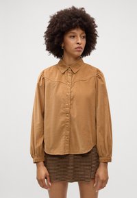 Camicia button-up in velluto a coste marrone con colletto classico, maniche lunghe a sbuffo e orlo arrotondato. Presenta una texture sottile e bottoni dorati.
