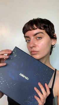 Mörkblå Zalando-låda med texturerad yta, guldfärgad logotyp och spridda guldfärgade fläckar, hålls av en person iklädd svart linne.