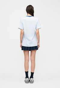 Femme debout, vue de dos, portant une chemise à manches courtes bleu clair, une jupe plissée marine, des chaussettes noires et des baskets grises, sur un fond blanc.