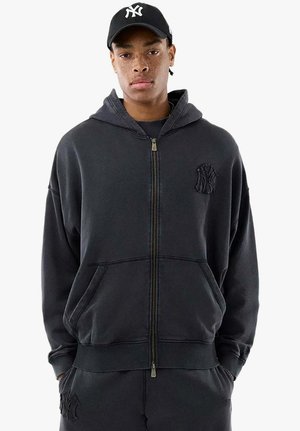 Giovane uomo che indossa una felpa nera con zip e pantaloni abbinati, con un cappellino nero dei New York Yankees, le mani in tasca, espressione neutra.