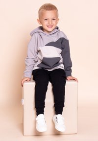 Uśmiechnięty młody chłopiec o blond włosach, ubrany w szarą i czarną bluzę z kapturem, czarne spodnie oraz białe trampki, siedzi na beżowej kostce na jednolitym tle.