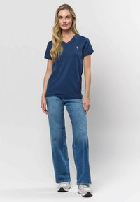 Tengerészkék V nyakú póló pamutkeverékből, világoskék, bő szárú farmerral párosítva. A mellkasán egy kis logó látható, és texturált anyagból készült.