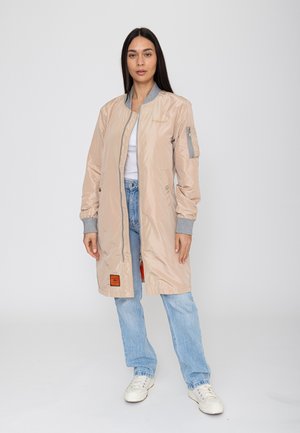 Frau steht frontal und trägt eine beige lange Bomberjacke, ein weißes Oberteil, blaue Jeans und weiße Sneakers vor einem einfarbigen hellen Hintergrund.