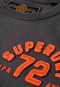 Camiseta de algodón gris con el texto bordado en relieve en naranja "SUPER 72" e "IPN", con un cuello de canalé y un acabado texturizado.