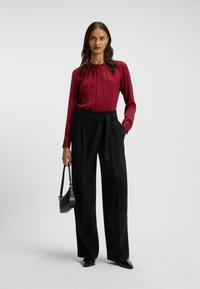 Blusa di seta bordeaux con pieghe, abbinata a pantaloni neri a vita alta e larghi. Il modello tiene una piccola borsa nera e indossa scarpe nere.
