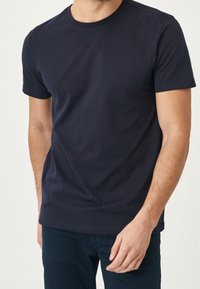 Uomo che indossa una maglietta a maniche corte blu scuro senza scritte e pantaloni scuri, in piedi contro uno sfondo neutro.