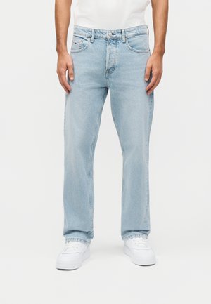 Man draagt lichtblauwe jeans met rechte pijpen, witte sneakers en een wit shirt, staand tegen een effen achtergrond.