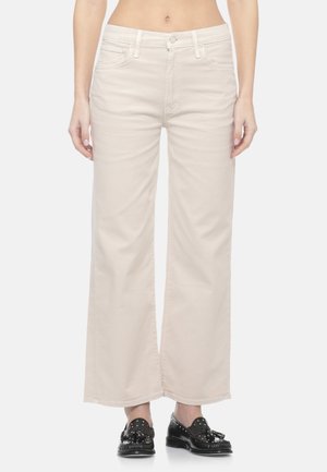 Donna che indossa pantaloni beige a vita alta e gamba larga e mocassini neri con nappine, in piedi con le braccia rilassate lungo i fianchi su sfondo bianco.