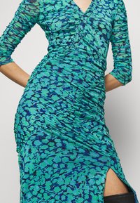 Robe portefeuille froncée bleu et verte avec manches trois-quarts et fente latérale dévoilant des bottes noires montantes.