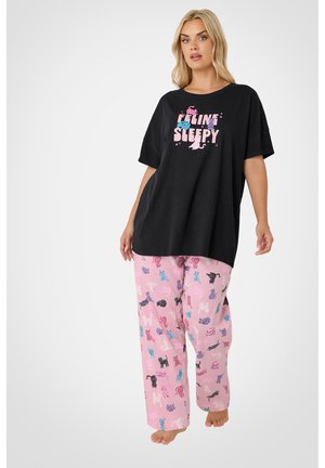 Kobieta ubrana w czarny oversize T-shirt z napisem „Feline Sleepy” oraz różowe spodnie do piżamy z kolorowymi nadrukami kotów, boso, stoi na białym tle.