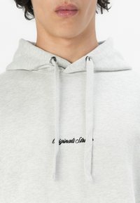 Felpa grigia chiara realizzata in un tessuto morbido, con un logo frontale in corsivo nero. Presenta lacci e occhiali metallici argentati per il cappuccio.