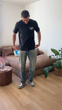 Giovane uomo con capelli intrecciati sistema una maglietta nera Rip Curl, indossa jeans azzurri e sneakers, in piedi in un soggiorno moderno con piante.