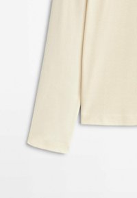 Manica lunga di un top in maglia a coste color crema con cuciture semplici sul bordo, su uno sfondo bianco.