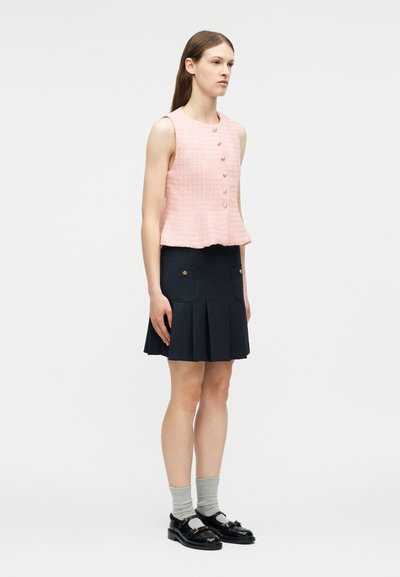 Γυναίκα που φοράει ροζ αμάνικο υφασμάτινο τοπ με κουμπιά, σκούρο μπλε πλισέ φούστα με τσέπες, γκρι κάλτσες και μαύρα loafers, στέκεται προφίλ.