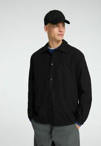 Giacca leggera nera con colletto, bottoni a pressione e polsini elasticizzati, abbinata a un maglione blu e un berretto nero, su uno sfondo neutro.