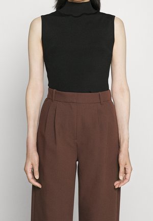 Top senza maniche nero con texture a coste e collo alto, abbinato a pantaloni marroni a vita alta con pieghe e tasche laterali.