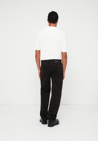 Chemise tricotée blanche à manches courtes, associée à un pantalon ample marron foncé et des chaussures noires, vue de dos.