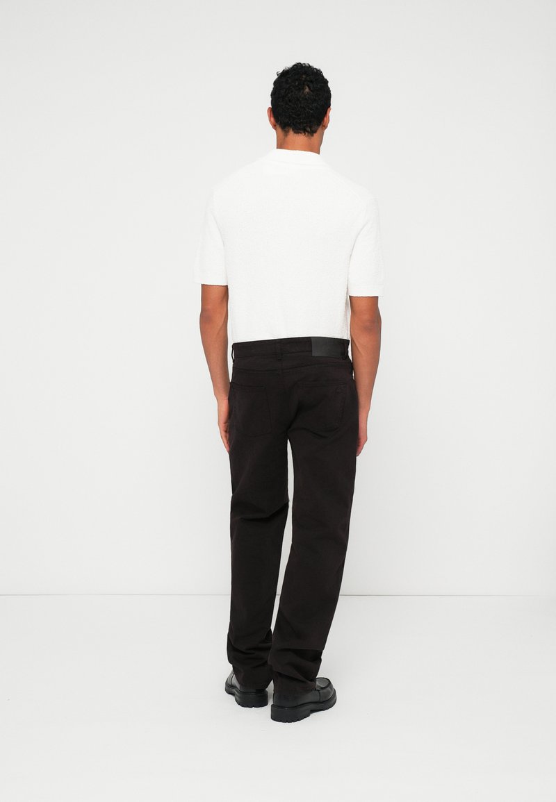 Maglietta bianca a maniche corte in maglia tessuta abbinata a pantaloni loose-fit marrone scuro e scarpe nere, vista da dietro.