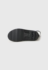 Semelle de chaussure noire avec des motifs de texture et des marquages, posée à plat sur une surface blanche.